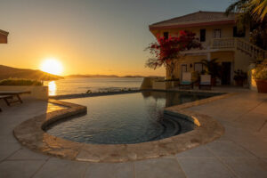 Swimming pool and stone patio outside A Dream Come True Villa with the sun setting on Mahoe Bay in the background.