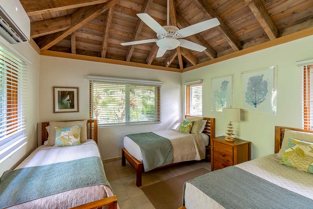 Beachcomber Villa guest room with three twin beds, lofted wood ceiling with a fan and windows looking out on tropical gardens.