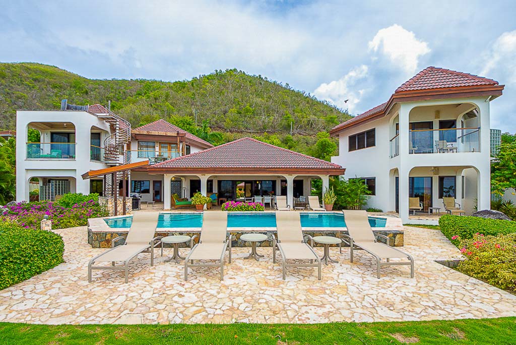 Caribbean Wind Villa with a white exterior and red tiled roofs in front of a green hill with a pool and patio in the foreground.