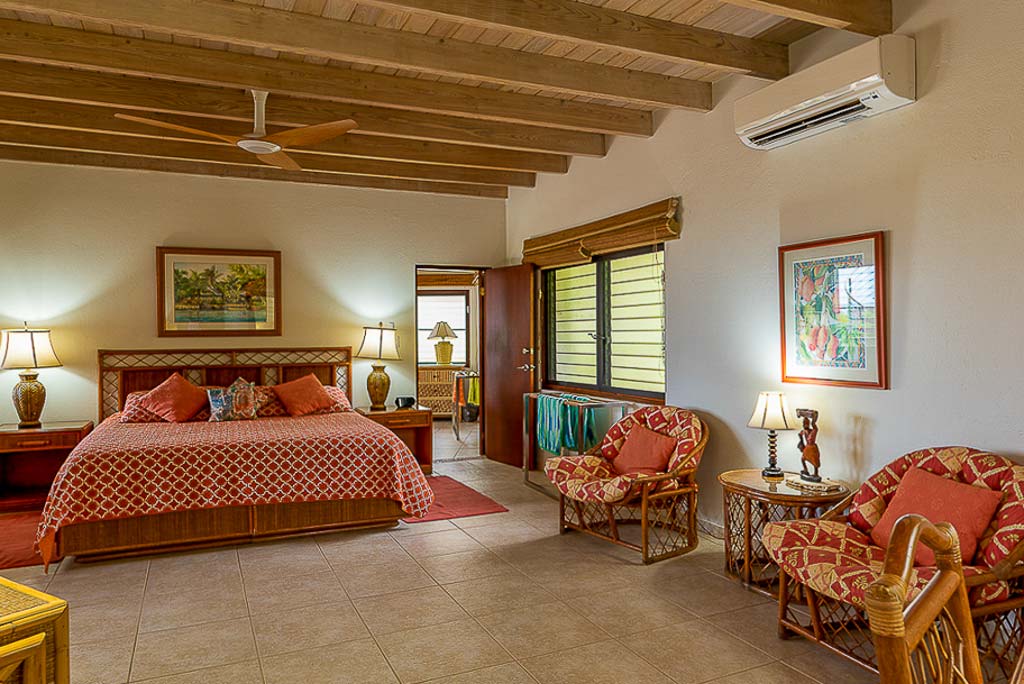 Spacious guest room with a bed and sitting area with tile floors, a wood-beam ceiling and a door open to a en-suite bathroom.