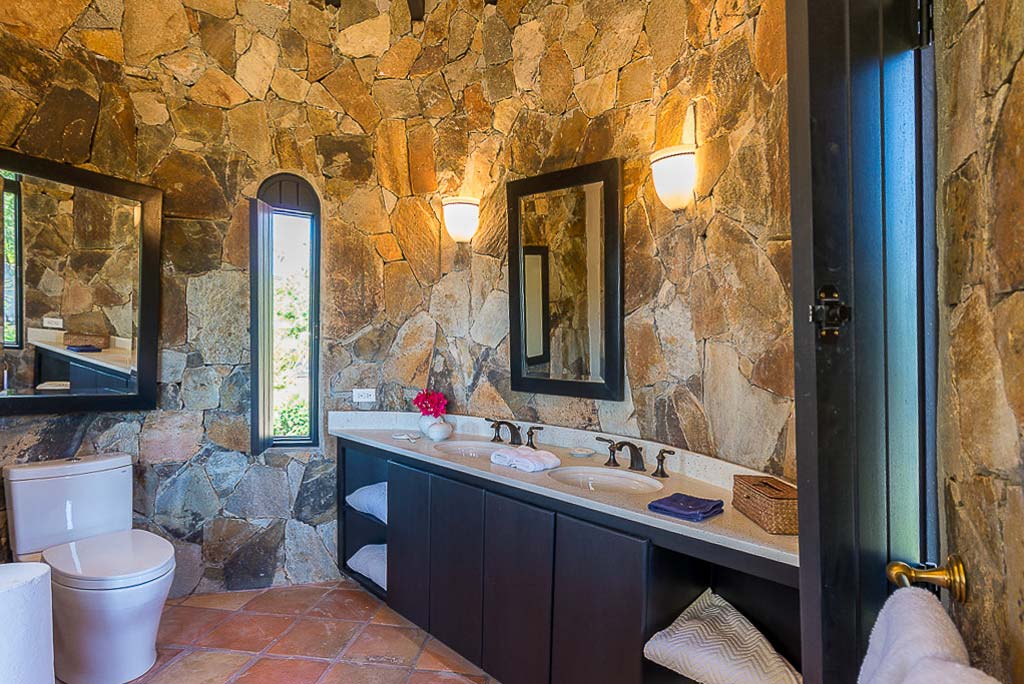 Turret-style double master bathroom with natural stone walls and tile floors and light coming in from an open window.