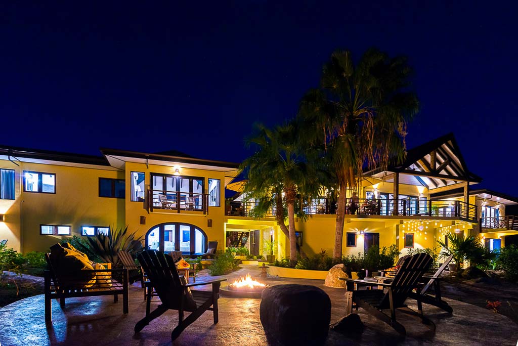 A roaring fire pit at night outside the yellow facade of the two-level Cornucopia Villa lit up by exterior lights.