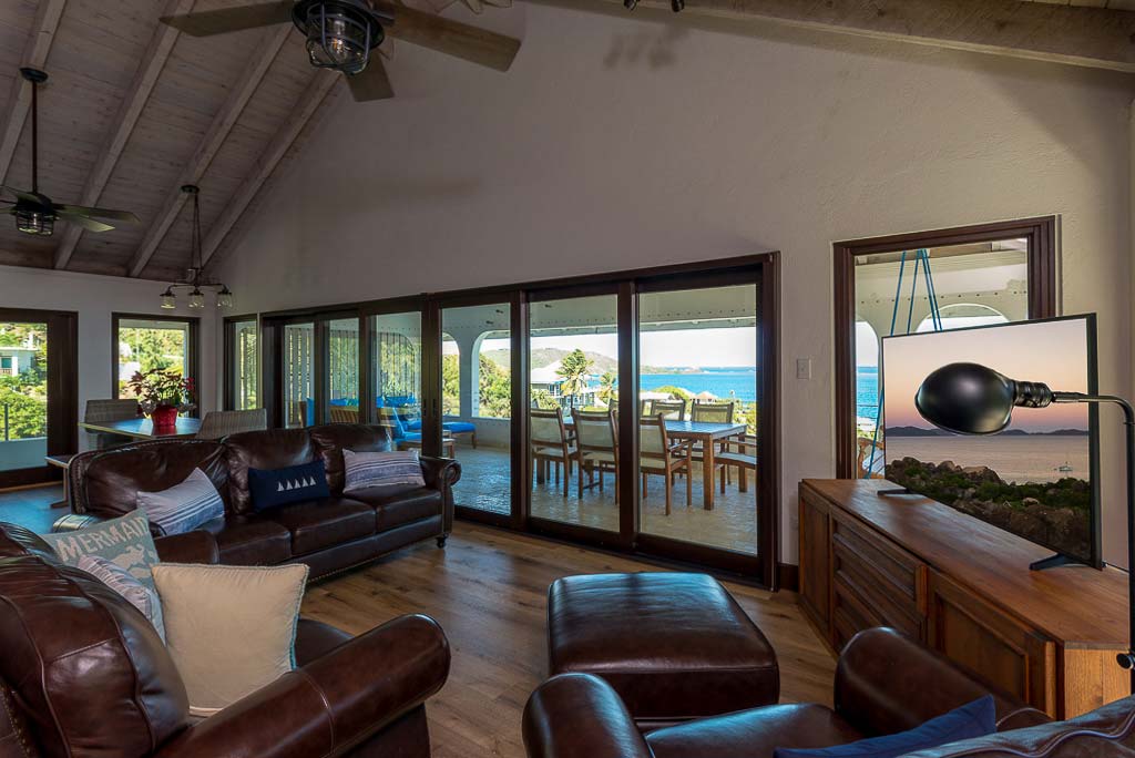 The living room at Dos Sols Villa with leather couches, a flat-screen TV and windows looking out on the deck and sea.