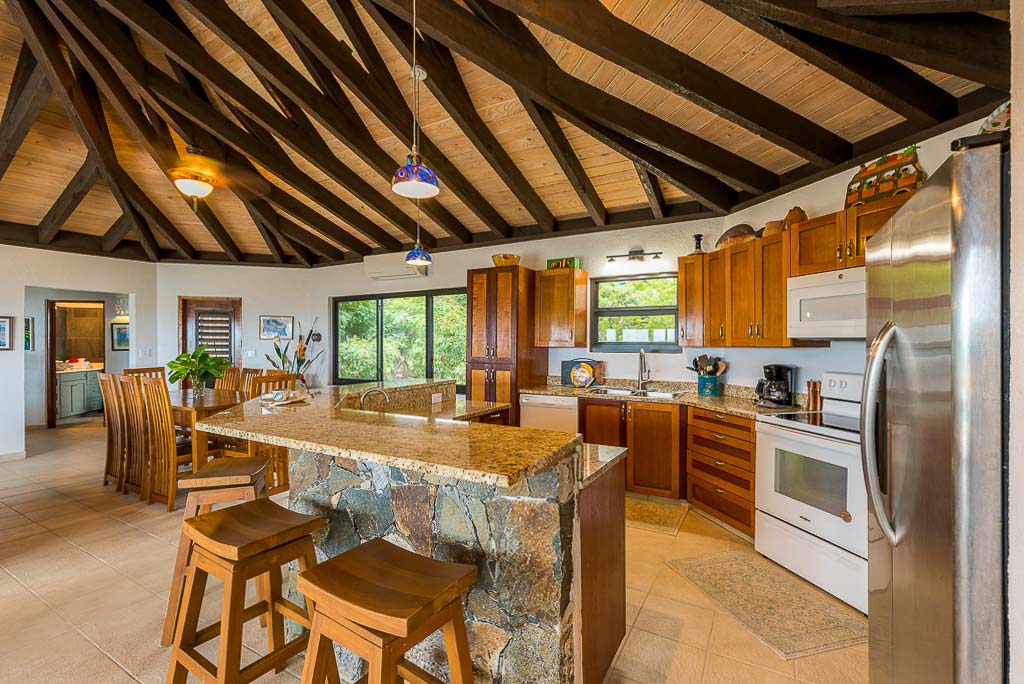 phoria villa’s kitchen with an island with a granite countertop and stools and a formal dining area in the background.