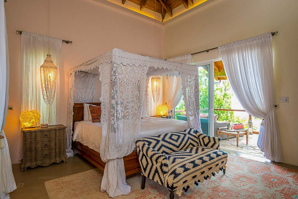 Master bedroom at La Vida Villa with a king-side four-post bed, white curtains and natural light from an open door to a patio.
