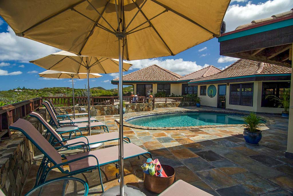 Large stone patio at Mon Repos Villa with a line of lounge chairs with umbrellas and a pool and the villa in the background.