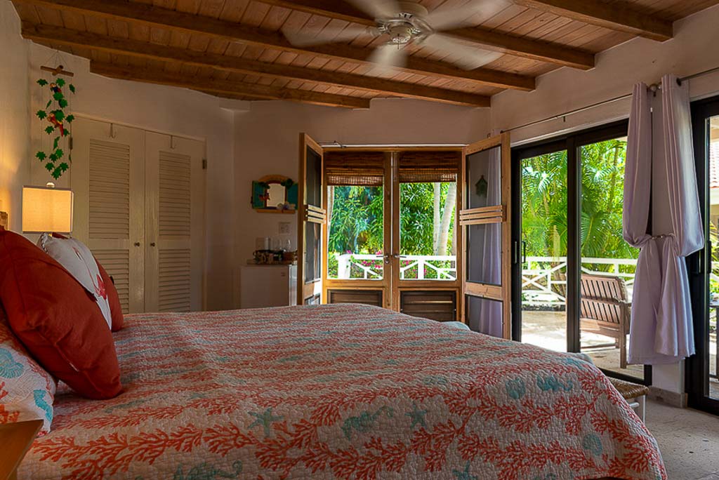 Sea Fans guest room with a king bed, wood-beam ceiling and glass doors out to a patio with palm trees in the background.