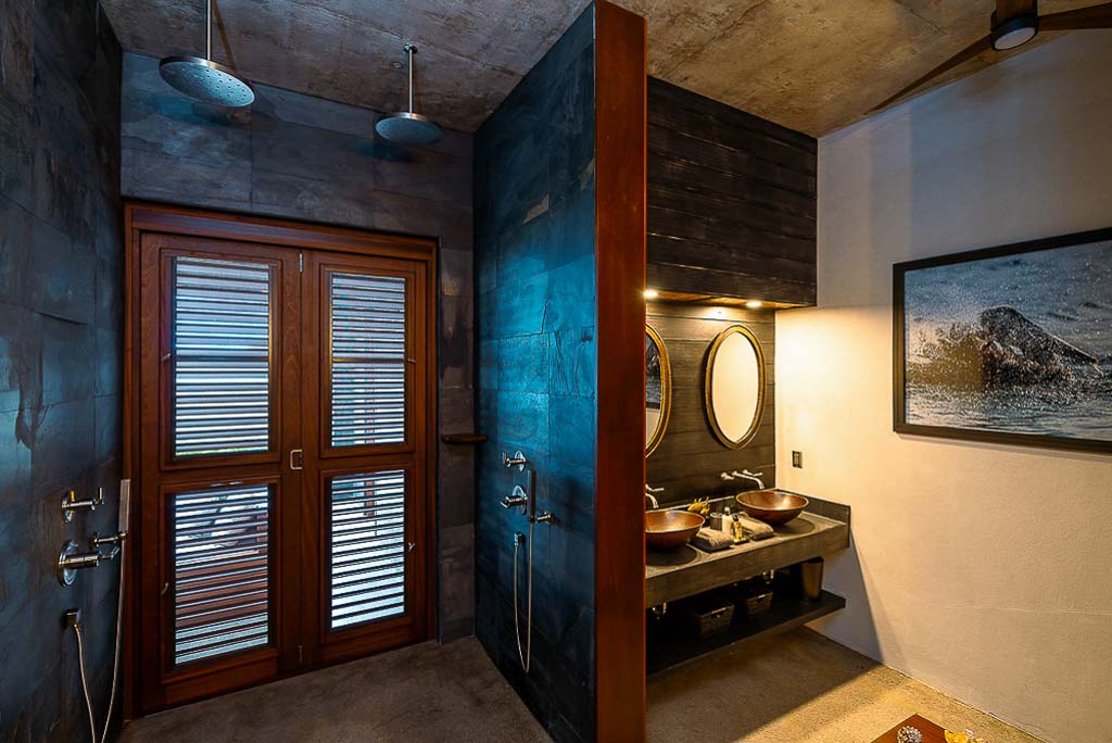 Segura Villa master bathroom with a double indoor/outdoor shower, double vanity and dark wood and natural stone walls.