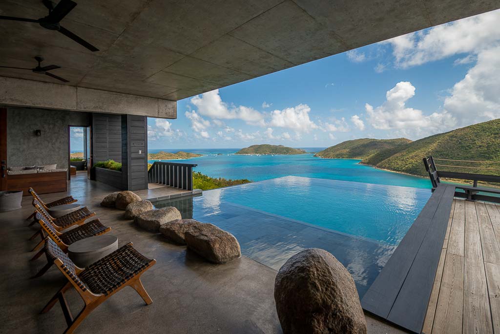 Natural stone open-air living space next to an infinity-edge pool with a backdrop of crystal blue Leverick Bay on a sunny day.