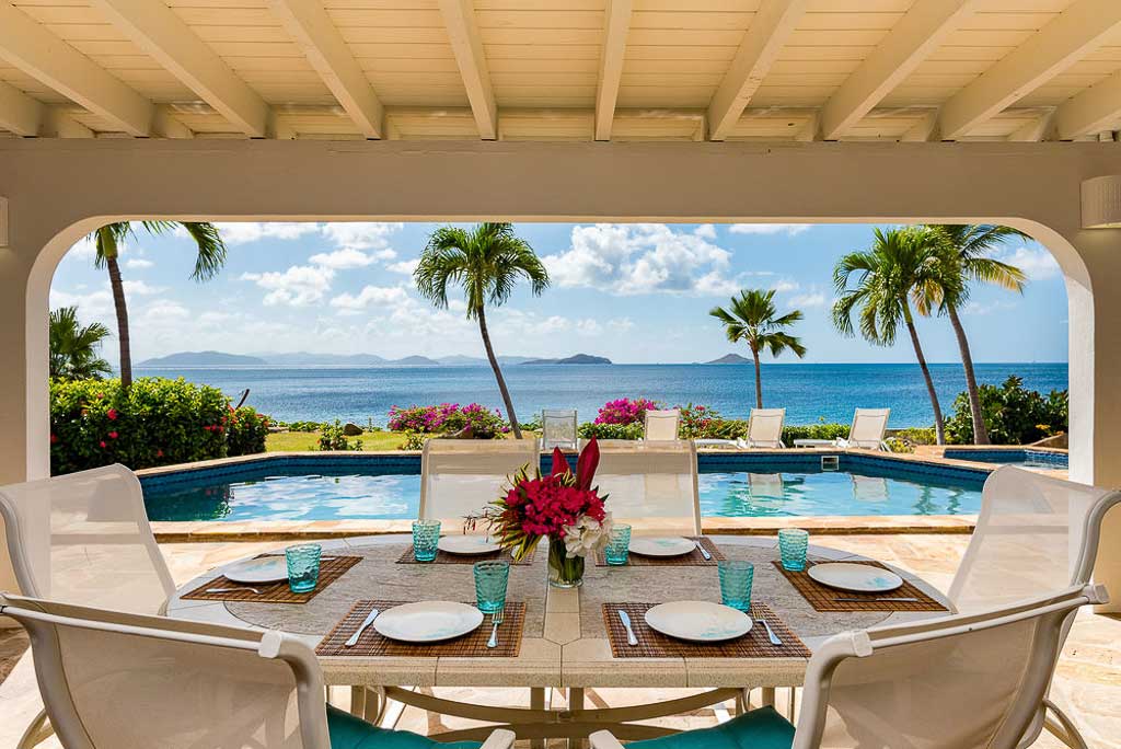 Beach Dreams Villa outdoor dining area on a covered pool-side patio with palm trees and Mahoe Bay in the background.