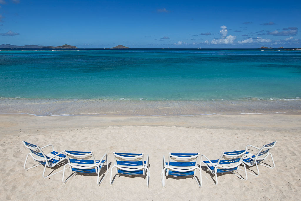Row of six beach chairs on a pristine white sand beach at Mahoe Bay facing the calm blue sea and sunny blue sky.