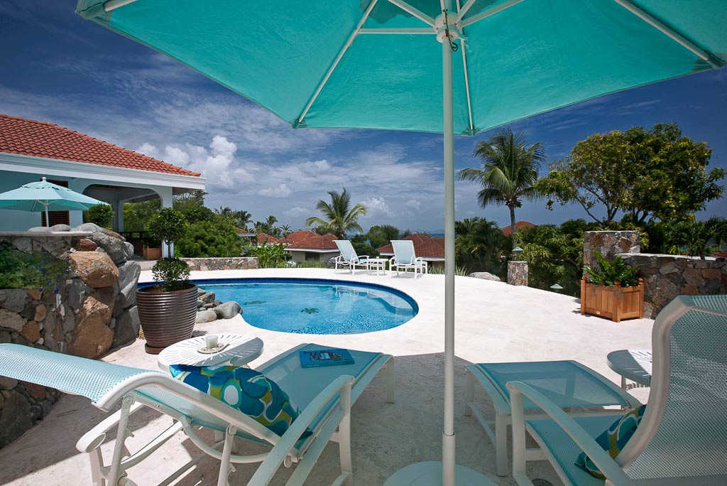 Lounge chairs under an umbrella next to a pool and Blue Lagoon villa with palm trees and a blue sky in the background.