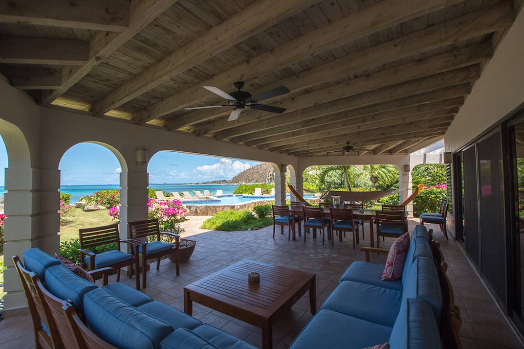 Spacious covered patio with separate dining and living areas and ceiling fans with a pool and Mahoe Bay in the background.