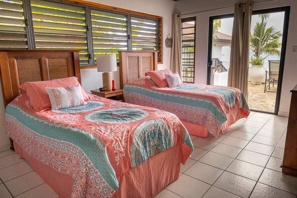 Guest room at Villa ValMarc with twin beds and tile floors and a glass door leading to a stone patio looking out on the sea.