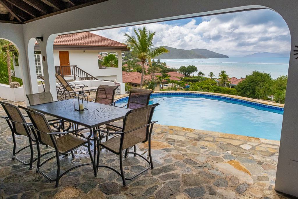 Covered poolside natural-stone patio with dining table at Villa ValMarc with the blue waters of Mahoe Bay in the background.