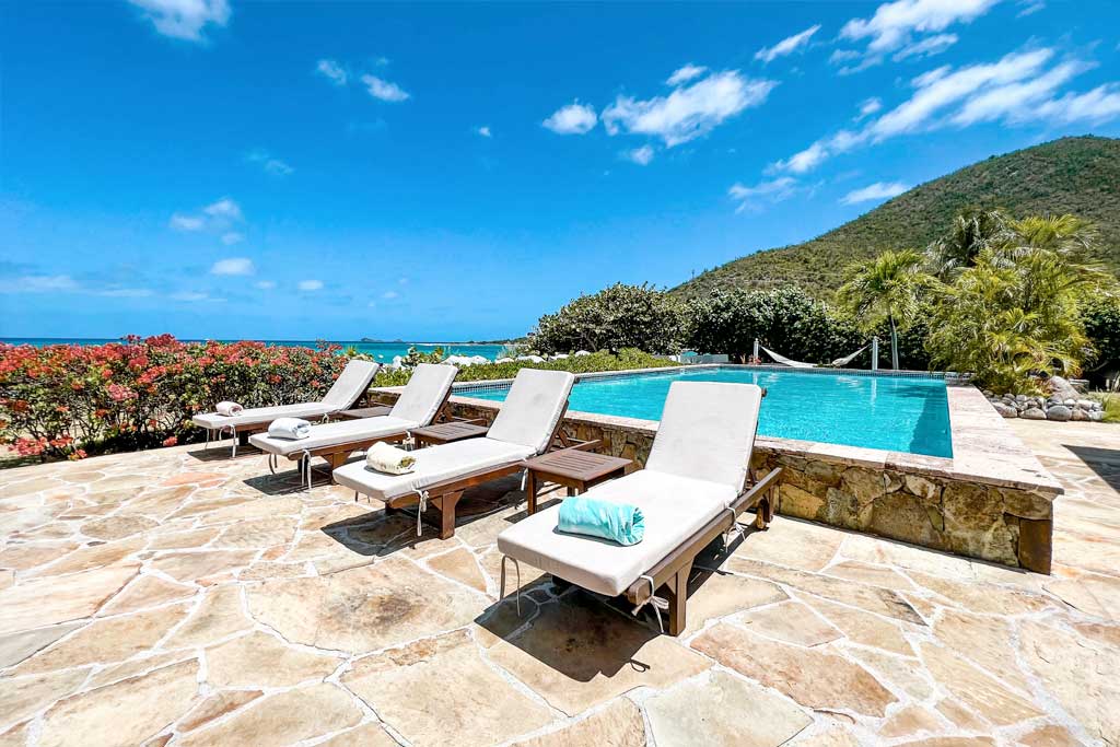 Row of lounge chairs and side tables next to a swimming pool at Sea Fans Villa with a backdrop of ocean and green hillside.