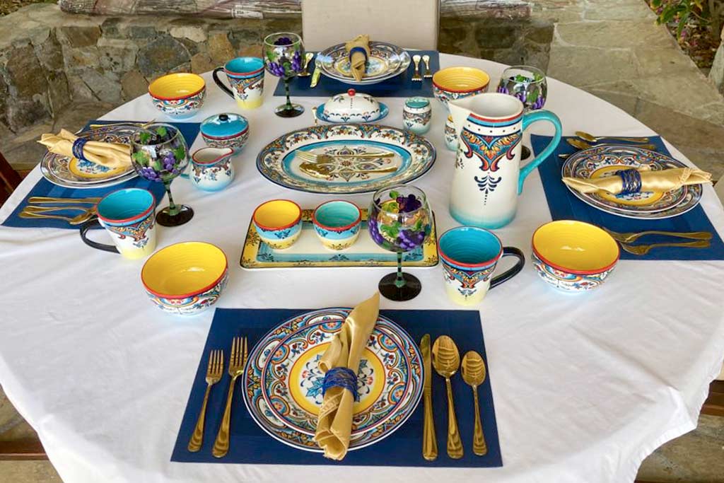 Round table set for a formal dinner with a white tablecloth and brightly colored plates and glasses on a natural stone patio.