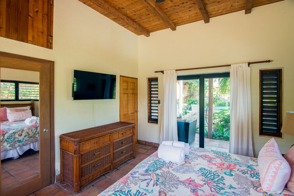 Ground-floor guest room with a king bed, stone-tile floor, wood bureau, mounted flat-screen TV and glass door to a patio.